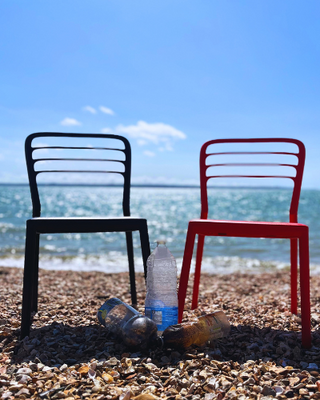 Two chairs, one black and one red, made from recycled plastics, placed on a pebble beach with the ocean in the background.