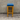 Wooden bar stool with a blue cushioned seat against a gray brick wall.