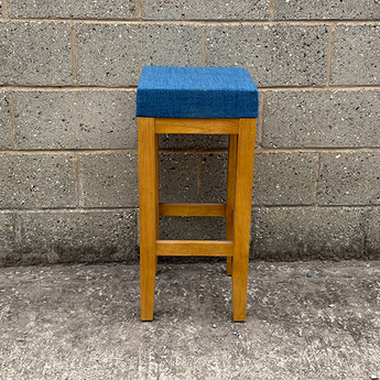 Wooden bar stool with a blue cushioned seat against a gray brick wall.