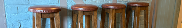 Four wooden stools with leather seats against a blue brick wall.